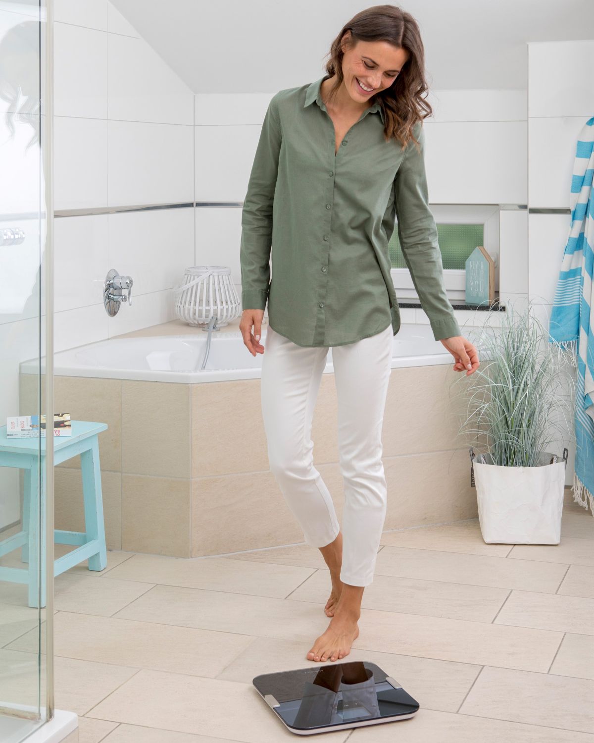 A smiling woman stands barefoot on a black digital body analysis scale in her bathroom.